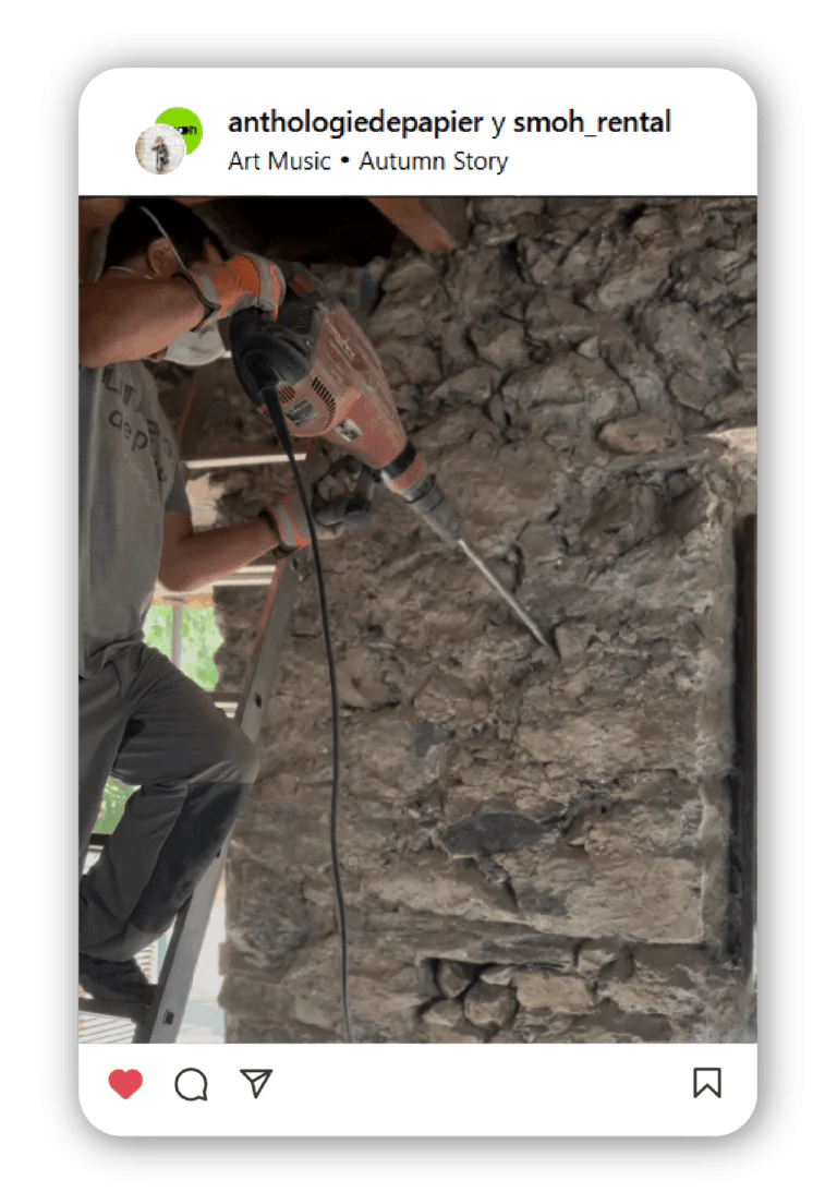 Worker wearing gloves uses an orange and black angle grinder on a rough stone wall, standing on a ladder at a construction site.