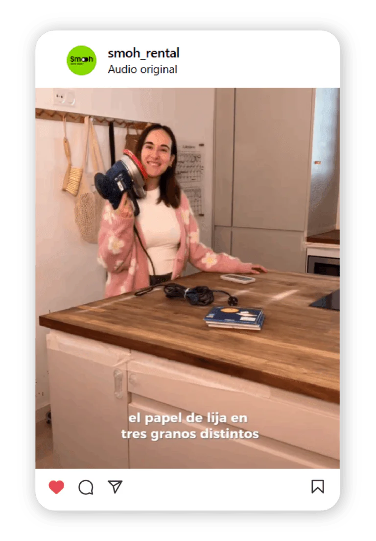 Smiling woman in a pink cardigan stands at a kitchen island, holding a large blue appliance near her shoulder while a phone sits on the counter in front of her.