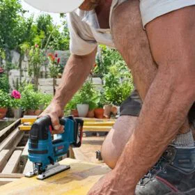 Construction worker in a white hard hat cutting a wooden plank with a blue cordless jigsaw on an outdoor deck.