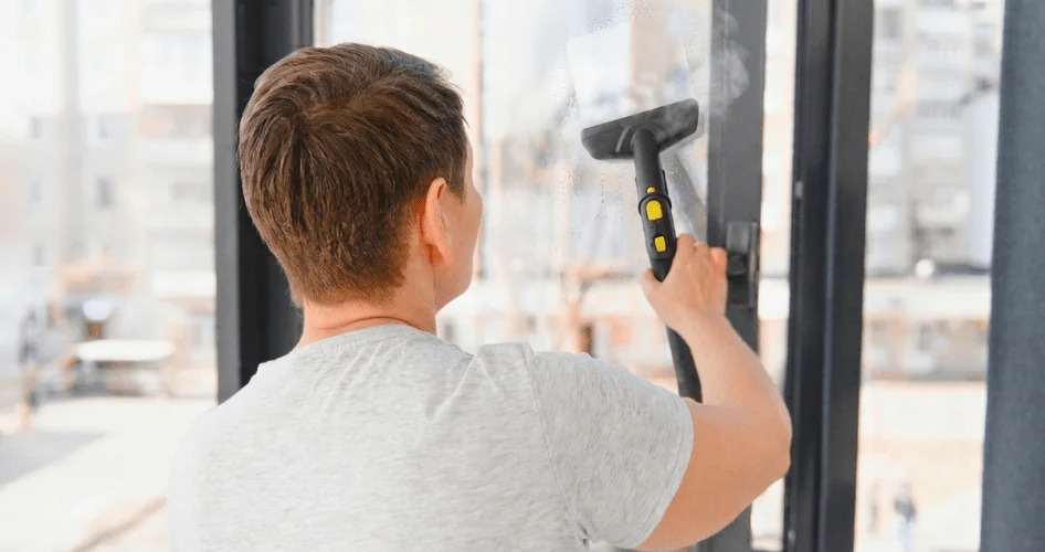Man in a gray t-shirt cleans a large glass storefront window with a squeegee, seen from behind.