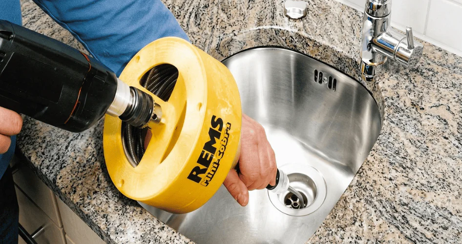 Plumber uses a yellow REMS tool attached to a drill to unclog a kitchen sink drain.